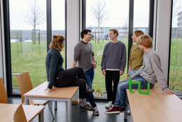 Group picture of students in a seminar room with the TU Dortmund logo. They speak together and look at each other. 